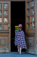 a girl in a pink cap and a large plaid cowboy shirt draped over her shoulders enters the doorway with her back to the camera through large beautiful doors with graffiti