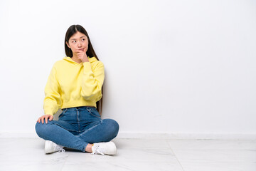 Young Chinese woman sitting on the floor isolated on white wall having doubts and with confuse face expression