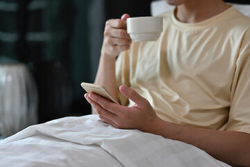 Close up of Asian male waking up on bed, checking a news and a social media while drinking a coffee in the morning.