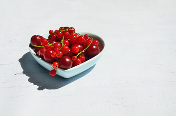 Red currants and cherries close-up in a heart-shaped ceramic vase on a textured light background. Minimalistic summer fruit idea with space for text.
