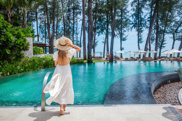 Travel woman walking near poolside in tropical resort, enjoy sea view. Female traveler in white...