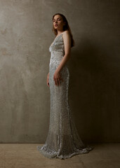 Fototapeta premium a slender woman with long red hair poses standing in a shiny silver dress looking at the camera with a confident expression on her face. Full-length photo