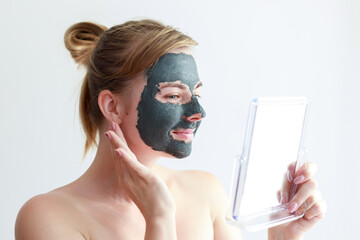 woman smiling in a cosmetic clay mask on her face looks in the mirror