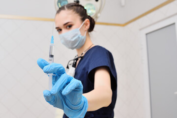 Fototapeta na wymiar a pretty young woman in a surgical suit in blue rubber gloves against the background of a surgical lamp holds a syringe in her hands.