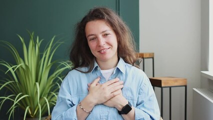 Smiling calm young caucasian woman holding folded hands on heart chest, breathing deep, feeling thankful at home.Showing gratitude, expressing appreciation, trust praying for peace, looking at camera