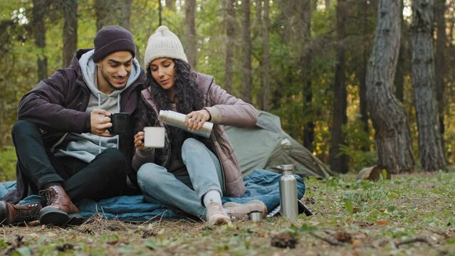 Young couple in love travelers sits in nature resting drinking tea from thermos enjoy hot drink resting talking smiling laughing feel pleasure travel enthusiasts on romantic date camping hike concept