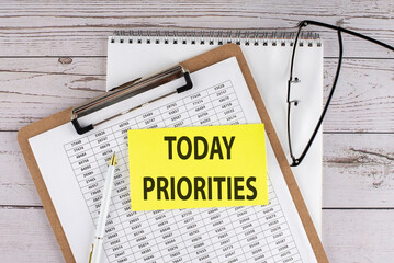 TODAY PRIORITIES text on yellow sticky on clipboard with chart, wooden background