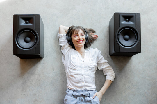 Young excited woman listening to the music in wireless headphones and sound speakers lying relaxed on concrete floor