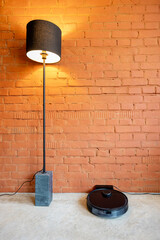 Robotic vacuum cleaner charging on dock station near floor lamp on brick wall background at home