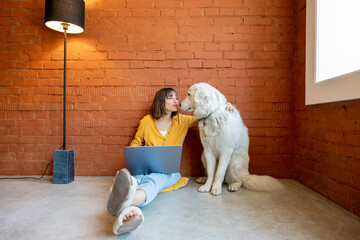 Young woman working on laptop computer while sitting with her cute dog on brick wall background at...