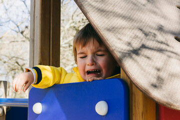 A little boy is crying while sitting on a slide in the playground.