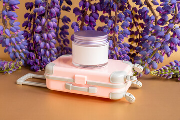 Moisturizing cream in glass jar with metal screw cap, small pink travel suitcase toy and purple lupine flowers a lot on brown background. Mockup, front view. Cosmetic moisturizer blank for branding.