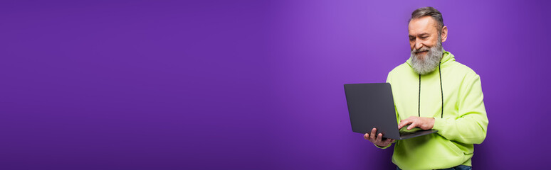 happy senior man with beard and grey hair using laptop on purple background, banner.