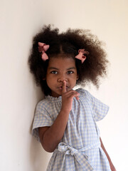 Cute little African girl standing against white wall saying hush be quiet or keep secret with finger on lips gesture.