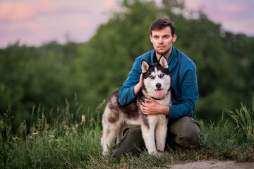 Naklejka na ściany i meble Portrait of a handsome young man and his pet Siberian Husky dog in nature in evening at sunset.