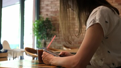 Woman, in a cafe, is waiting for an order, holding a mobile phone in her hand, typing, scrolling through the page using the application. Internet news app content concept of mobile phone dependency.