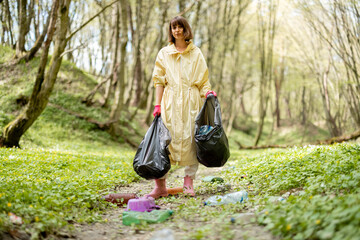 Young woman in protective clothes stands with garbage bags collecting scattered plastic in the woods. Problem of bed ecology and environmental pollution with long-decomposing waste