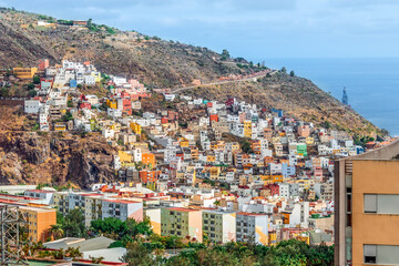 Village with multicolor houses at the foot of a mountain against the backdrop of the Atlantic Ocean in Santa Cruz de Tenerife, Spain. Colorful cityscape of the capital of the Canary Islands - obrazy, fototapety, plakaty