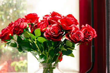 Bouquet of beautiful fresh scarlet red roses in glass vase stands on a table on a windowsill....