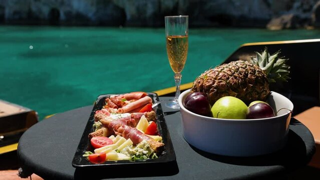 A set of meet and cheese plate, fruit and a glass of wine on the yacht deck. Man's hand take jamon from the table.