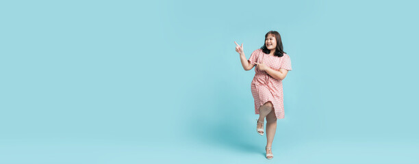 Fototapeta premium Full length Confidence cheerful beautiful Asian woman plus size in pink pastel dress with hands pointing to copy space aside on isolate banner light blue studio background.