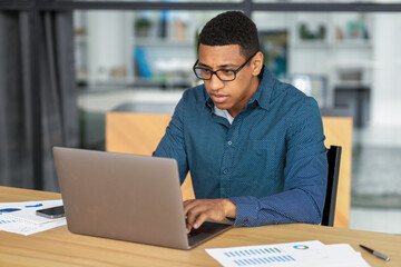 Concentrated African American businessman working from contemporary office. Young office worker man, looking at laptop screen, typing on keyboard while sitting at table