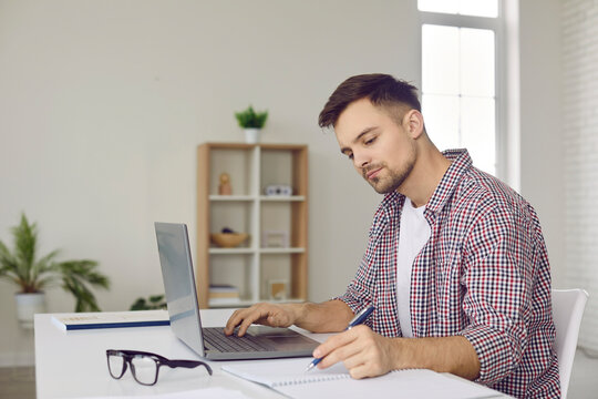 Remote University Student Taking Distance Classes. Focused Handsome Young Man Sitting At Desk At Home, Using Laptop Computer And Writing In Notebook. Remote Education, Studying Online Concept