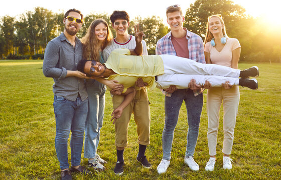 Portrait of funny young friends having fun, laughing and fooling around in park in summer. Five Caucasian people in casual clothes are holding their African American friend who is lying in their arms.