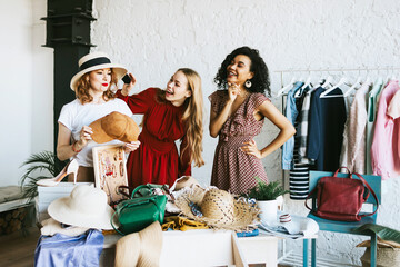 Obraz premium three young woman female caucasian and african students at swap party try on clothes, bags, shoes and accessories, change clothes with each other, second hand for things, zero waste life, eco-friendly