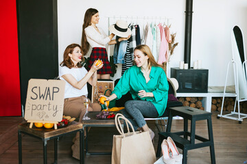 three young woman female caucasian students at swap party try on clothes, bags, shoes and accessories, change clothes with each other, second hand for things, zero waste life, eco-friendly