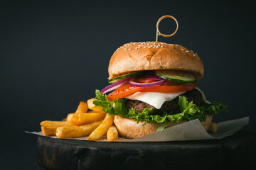 Big beef patty burger with cheese tomato purple onion cucumber and lettuce next to french fries on wooden black stand on black background