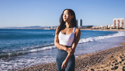 Slim Asian woman in grey leggings pants and white top looking away and dreaming about wellbeing vitality spending weekend at seashore beach near ocean, Korean fit girl reaching healthy lifestyle