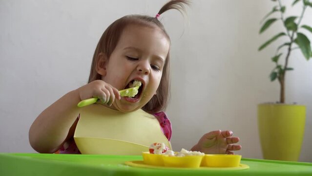 Close up of lovely baby girl eating have breakfast eggs vegetables sausage by herself at home. Adorable possitive happy toddler child eats with dirty face at table indoor. Food, nutrition healthy life