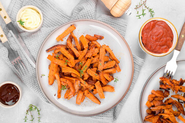 Delicious homemade roasted sweet potato with herbs, salt and pepper roasted in the pan on a ceramic plate and mayo, ketchup and barbeque sauce on a light grey table background, top view, above