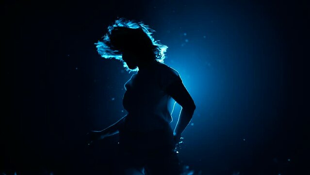 Young woman moves her head and hair in dark studio. Dancer performing dance on blue colorful spotlight background. Concept of sexual dancing with neon backlight.