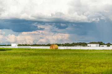 Obraz premium Hay roll bales on countryside field in Alachua in north Florida, USA rural area with farmland meadow and industrial white building in background and stormy sunset sky clouds