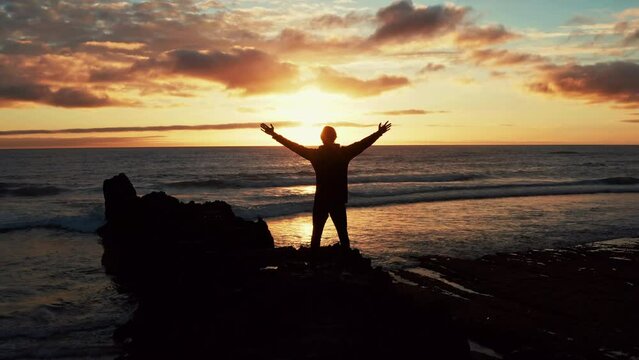 Man stands on a rock against the ocean  is looking distance during beautiful summer sunset. Human with raised hands looks to the sun over horizon in morning while sunrise. Freedom. Beauty of nature.