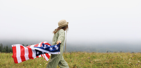 Beautiful child girl in hat with the American flag on foggy mountains. Independence Day of United...