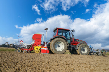 Fototapeta premium Agrarmotive - Frühjahrsbestellung. Landwirt mit rotem Schlepper und roter Drillmaschine bei der Aussaat von Sommergetreide in hügeliger Landschaft. 