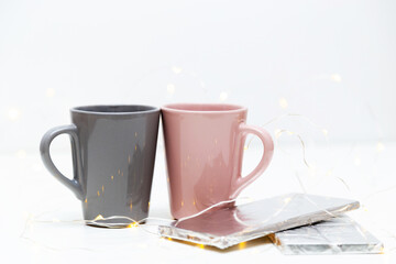 Two pink and grey coffee mugs mockup and a bar of chocolate on white background with small golden lights and bokeh. Blank template for your design, branding, business.