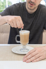 A young man in a cafe at a white table with a cup of latte coffee stirs with a spoon.