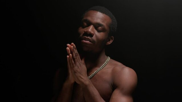 Portrait serious African American man experiencing onset of tragedy in life and praying with his hands clasped in front of him with clasped praying hands in dark studio with one light source on top.