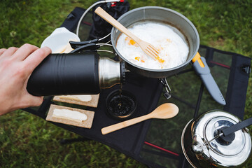 A tourist pours hot tea from a thermos into a mug, fried scrambled eggs in a frying pan on a hike,...