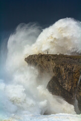 man walking along a cliff during a strong sea storm as a wave passes over him