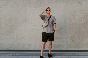 Portrait of handsome confident stylish hipster man in sunglasses with bag handy, standing guy near the concrete wall. Calm relaxing moment, summer vacation concept - 514533137