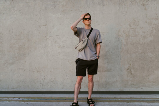 Portrait of handsome confident stylish hipster man in sunglasses with bag handy, standing guy near the concrete wall. Calm relaxing moment, summer vacation concept
