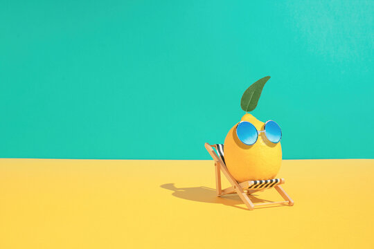 Lemon fruit chilling in beach chair on the blue and yellow background. Summer vacation concept. Sunglasses on lemon with green leaf relaxing on the sunbed. Creative art minimal aesthetic.