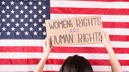 Woman is holding a sign Womens Rights Are Human Rights US flag on background. Protest against anti...