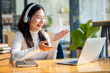 Cheerful and beautiful Asian woman wearing headphones and looking at digital tablet in video conferencing and online classes on the Internet at a coffee shop.