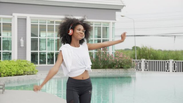 Young African American woman wearing headphones listening music standing dancing, female wearing earphones while enjoy with happiness at poolside, emotion and feeling, lifestyles concept.